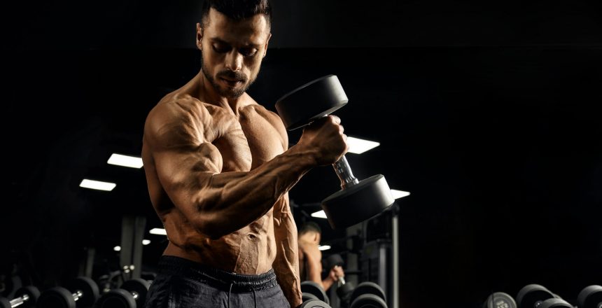 Side view of shirtless bodybuilder training biceps with dumbbell. Close up of muscular sportsman with perfect tensed muscular body posing in gym in dark atmosphere. Concept of bodybuilding.